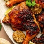 Grilled pollo asado with char marks on a white plate, garnished with cilantro and served with charred lime halves and tortillas.