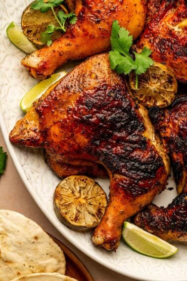 Grilled pollo asado with char marks on a white plate, garnished with cilantro and served with charred lime halves and tortillas.