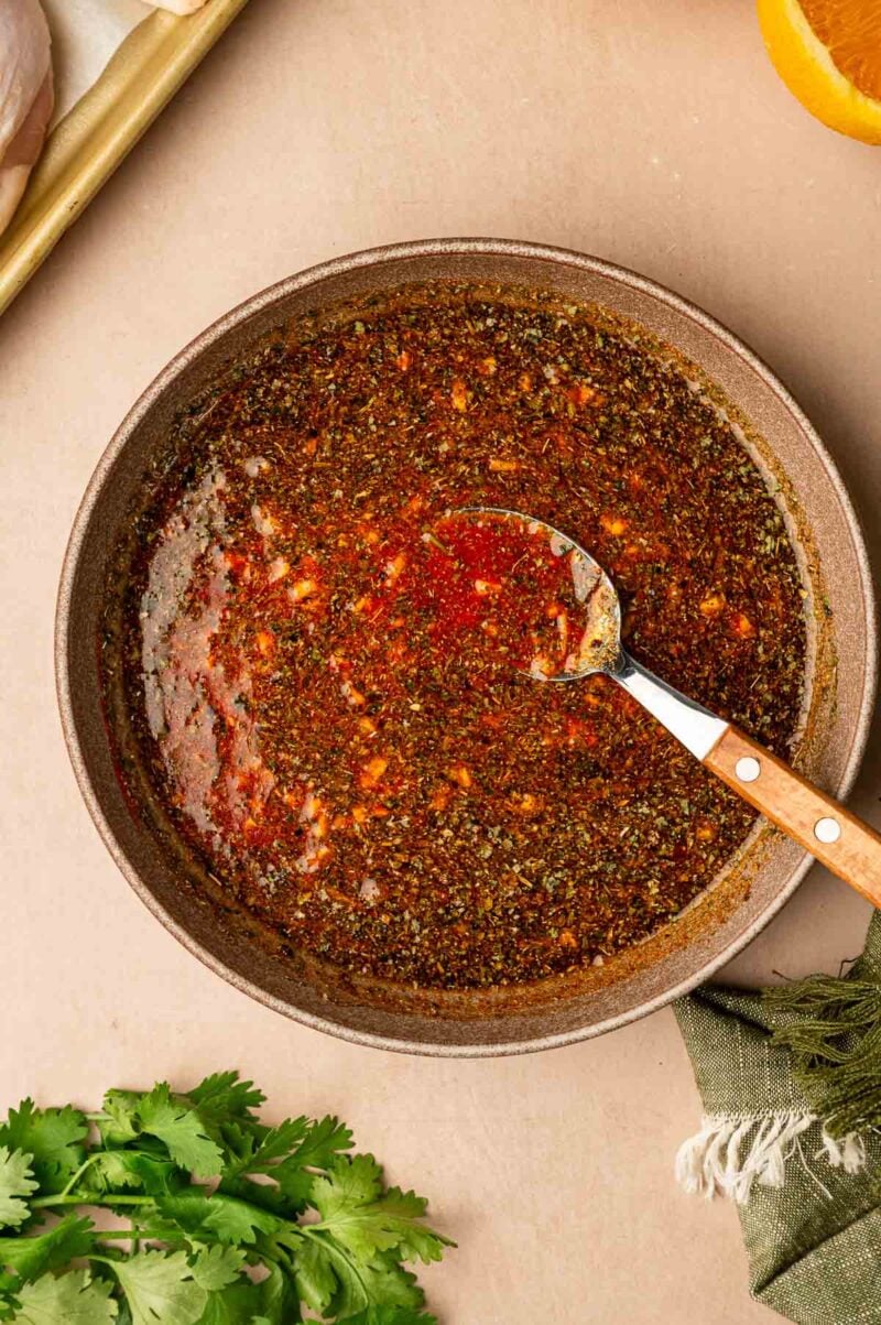 A bowl of seasoned marinade with a spoon resting inside, surrounded by fresh cilantro, a napkin, and part of an orange visible on the side.