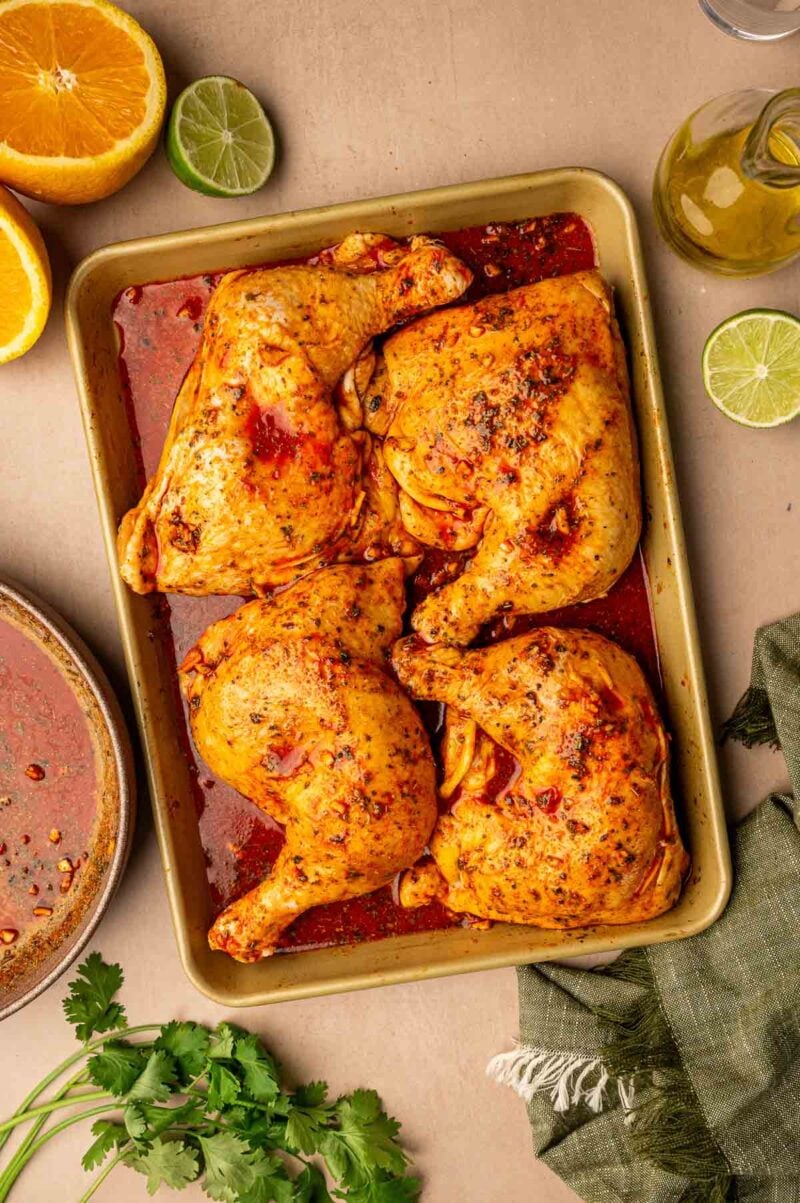 Four marinated chicken leg quarters on a baking tray, surrounded by cut citrus fruits, fresh cilantro, and a green napkin.