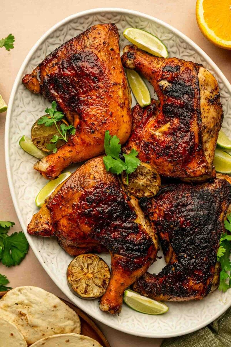 Four grilled chicken quarters on a white oval platter, garnished with lime wedges, cilantro, and grilled lime slices.