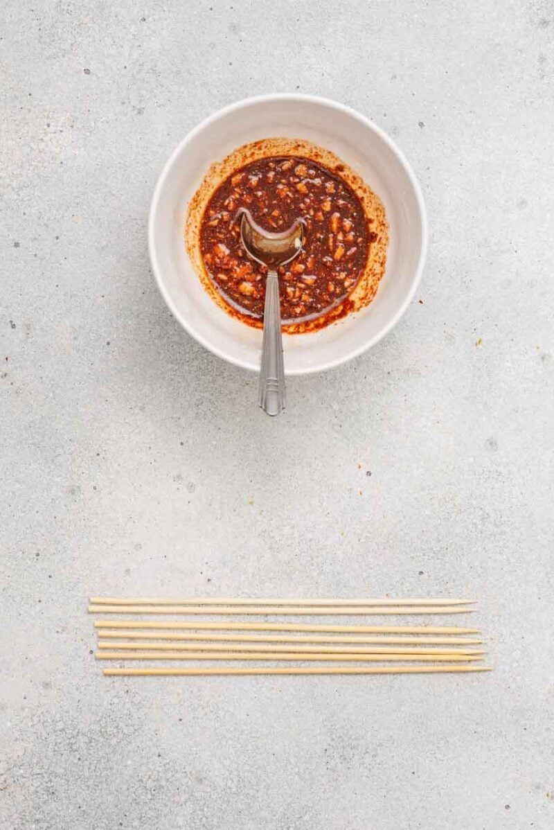 A white bowl of chili oil with a spoon inside is placed above a row of wooden skewers on a light gray surface.