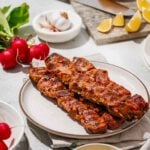 Grilled skewers of marinated meat served on a plate with mustard sauce, radishes, garlic, and lemon wedges on a table.