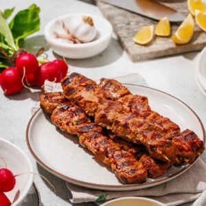Grilled skewers of marinated meat served on a plate with mustard sauce, radishes, garlic, and lemon wedges on a table.