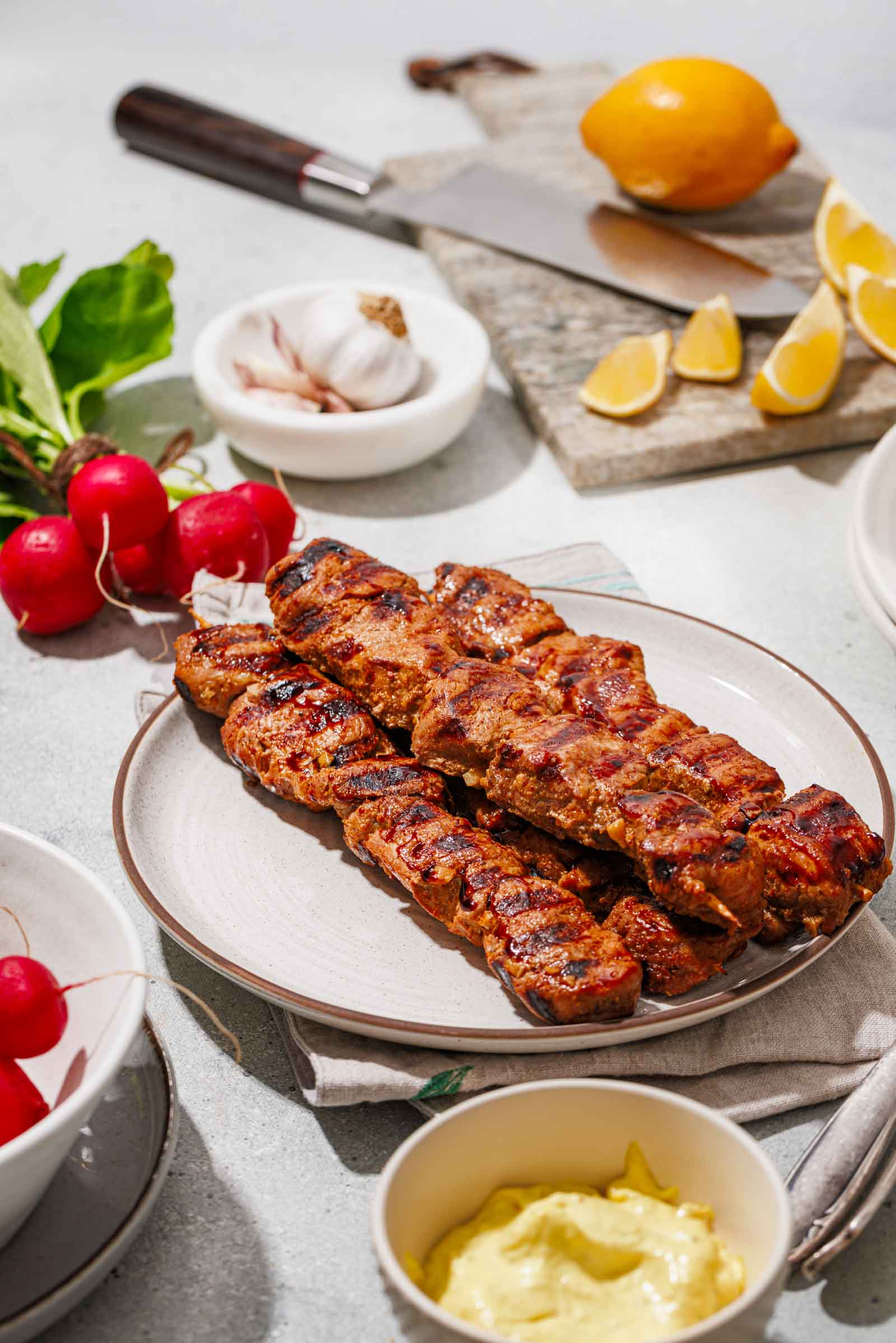 Grilled skewers of marinated meat served on a plate with mustard sauce, radishes, garlic, and lemon wedges on a table.