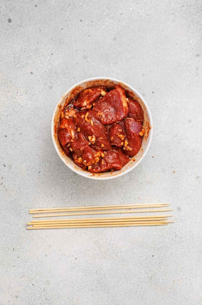 A bowl of marinated meat sits above five wooden skewers on a gray, textured surface.