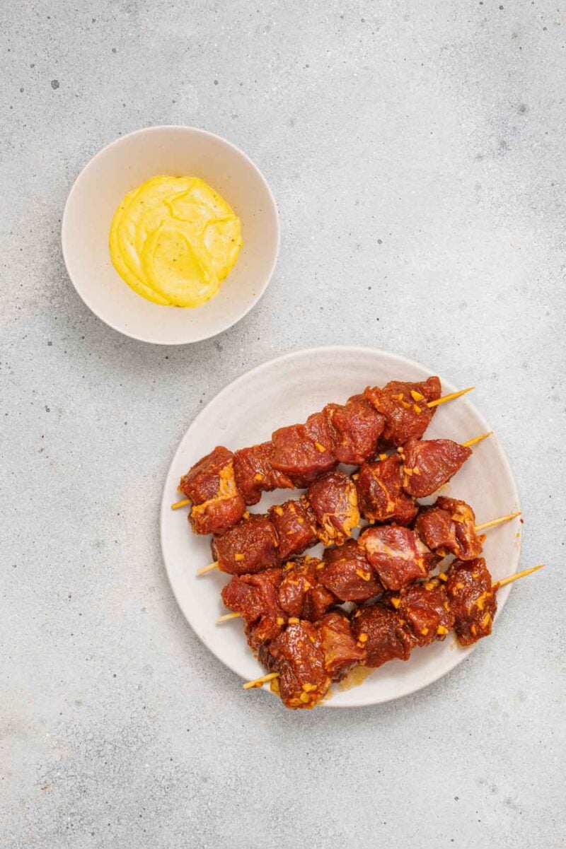 A plate of skewered, marinated meat sits next to a bowl of yellow sauce on a light, textured surface.