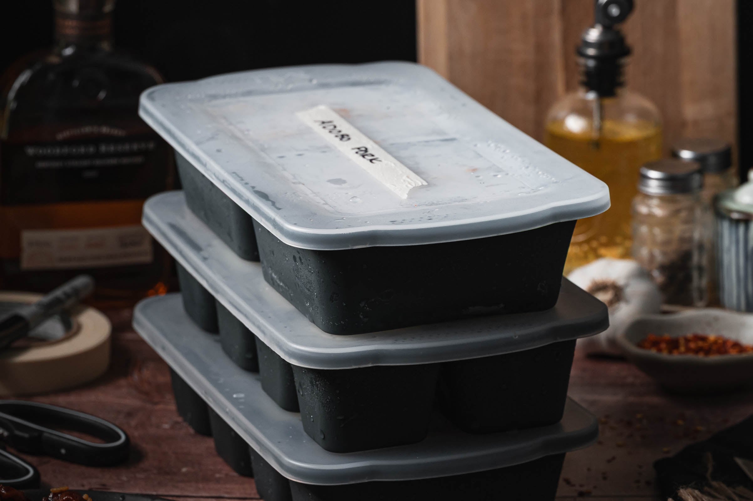 Three stacked black freezer meal prep trays with translucent lids sit on a wooden surface, surrounded by kitchen items such as a bottle, oil dispenser, scissors, and spices.