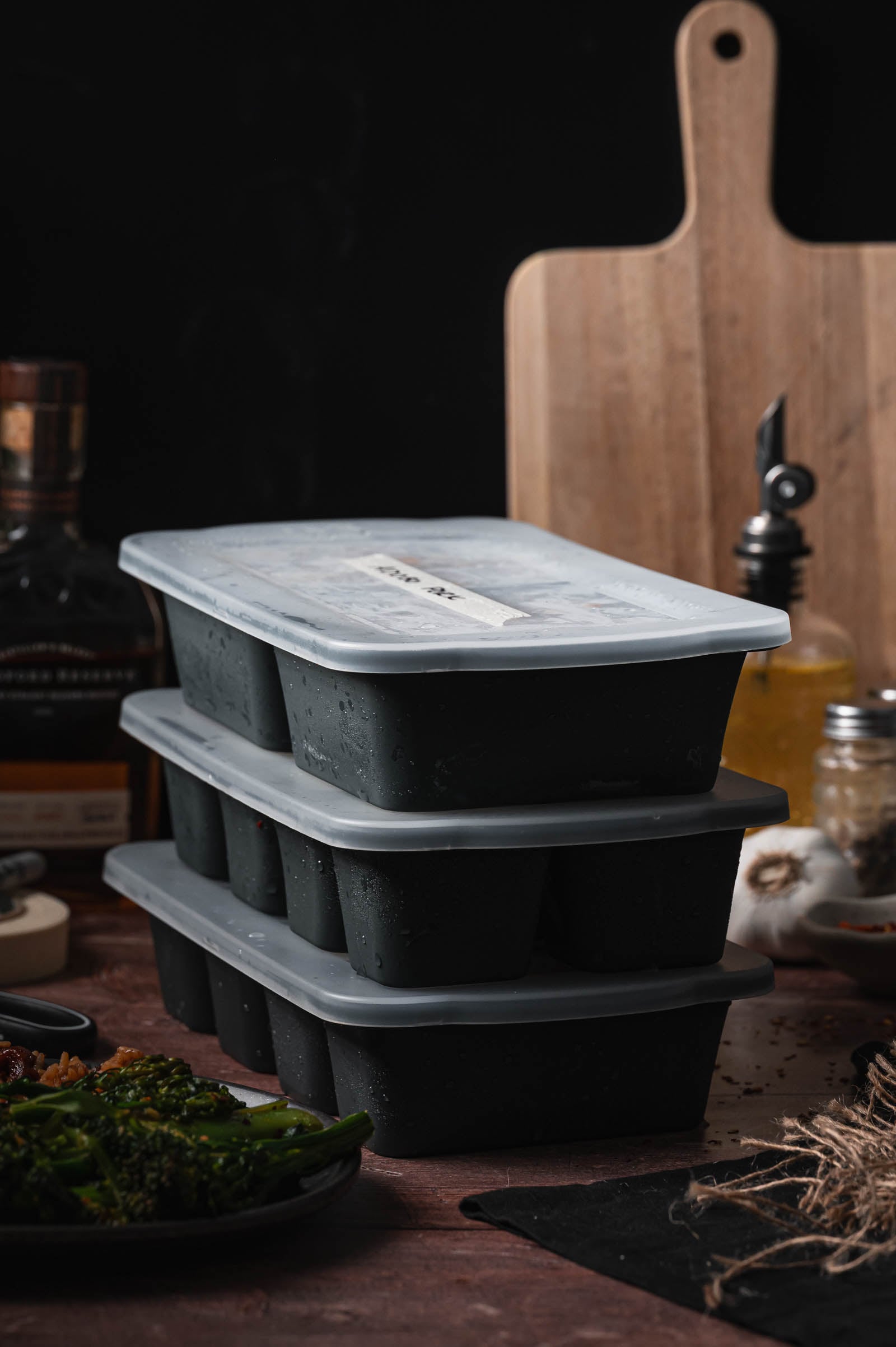 Three black meal prep containers with gray lids are stacked on a kitchen counter, with cooking ingredients and utensils in the background.