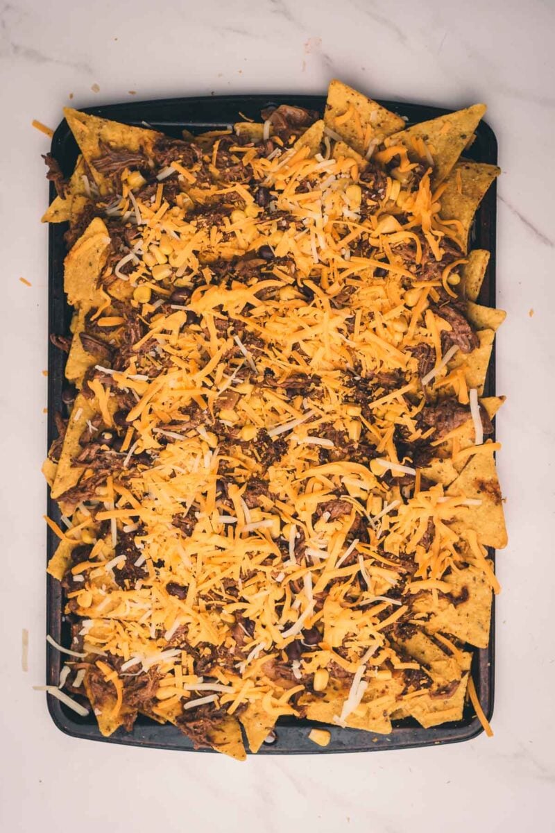A baking sheet filled with tortilla chips topped with shredded cheese and shredded beef, ready to be baked for nachos.