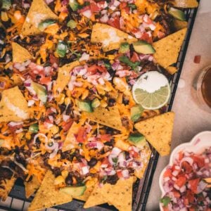 A tray of nachos topped with melted cheese, beans, corn, diced tomatoes, onions, avocado, and a lime slice, with a bowl of salsa and sliced avocado nearby.