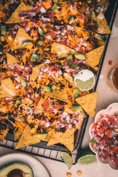 A tray of nachos topped with melted cheese, beans, corn, diced tomatoes, onions, avocado, and a lime slice, with a bowl of salsa and sliced avocado nearby.