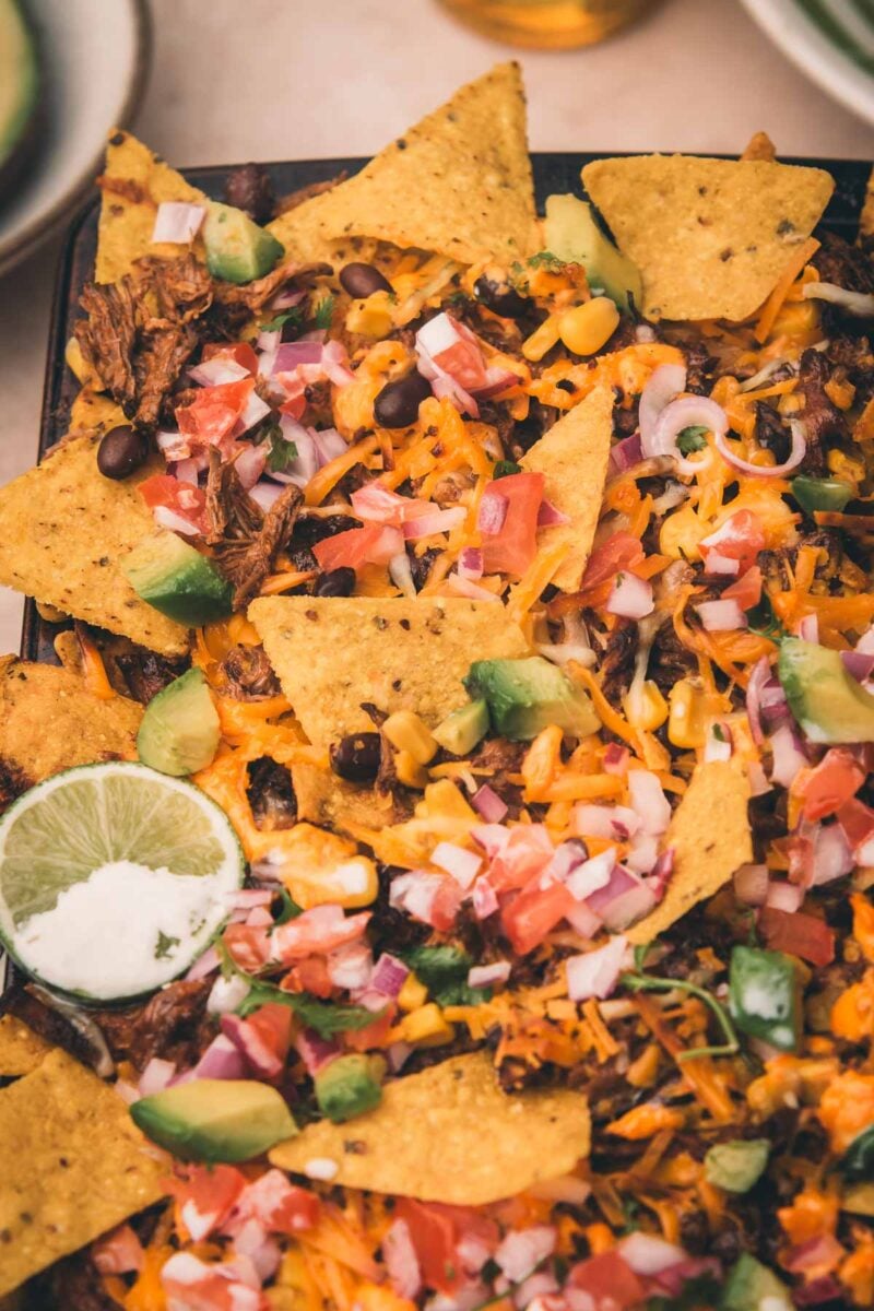Nachos topped with melted cheese, shredded Mexican beef, black beans, avocado, diced tomatoes, red onions, corn, and a slice of lime.