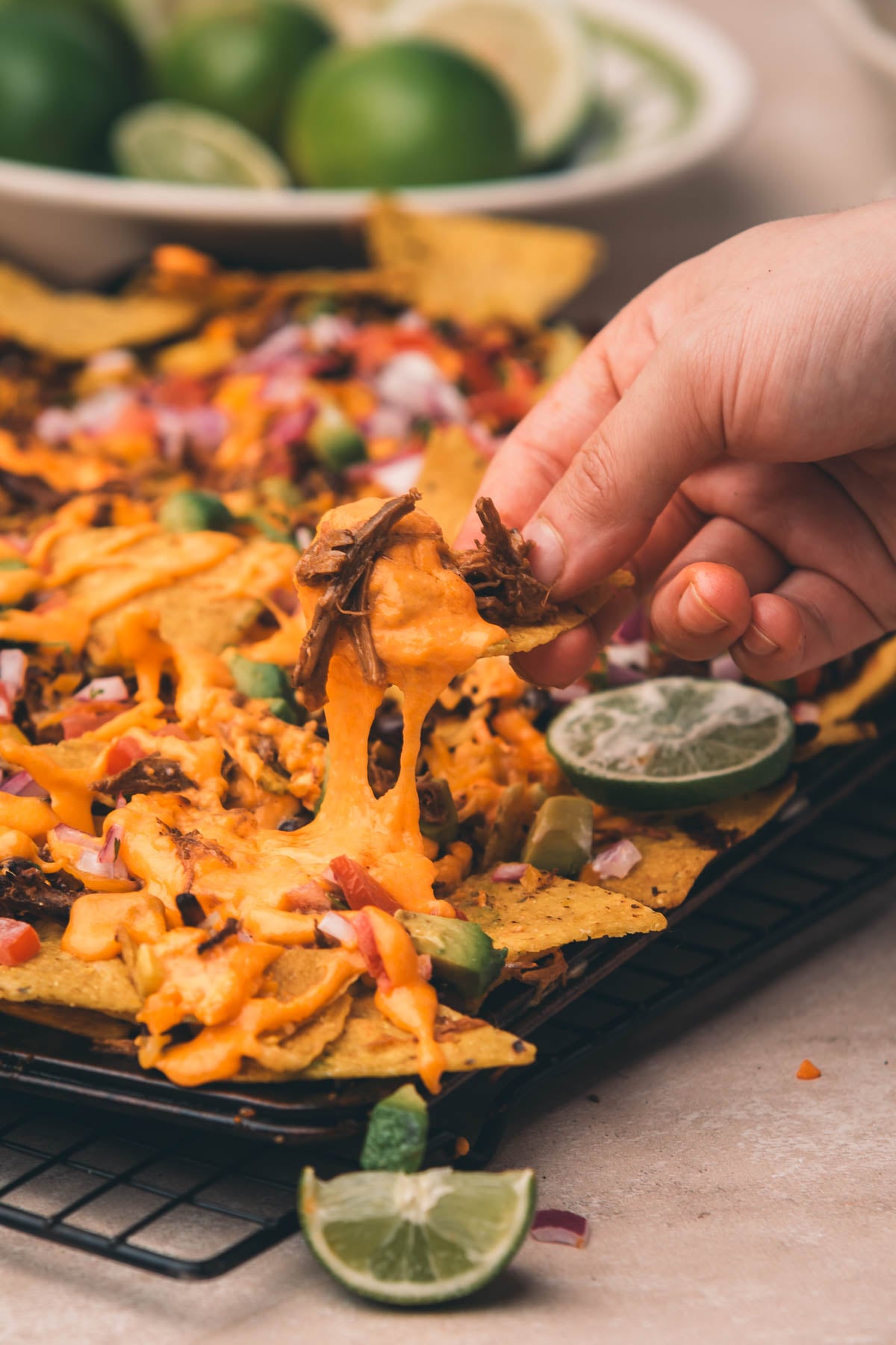 A hand lifts a cheesy nacho from a tray topped with melted cheese, jalapeños, and shredded meat. Lime wedges are visible in the foreground and background.