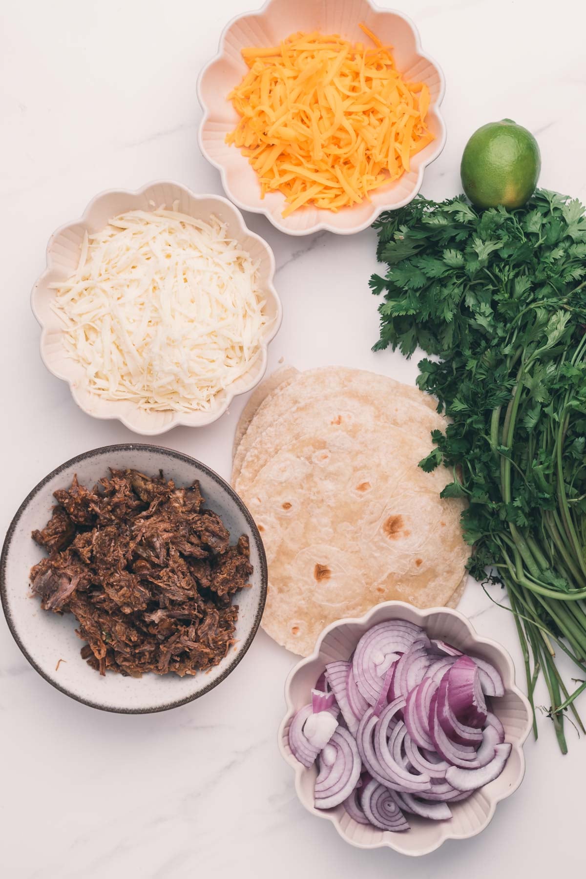 Shredded cheeses, sliced red onions, and shredded beef; flour tortillas, cilantro, and a lime are also on a white surface.