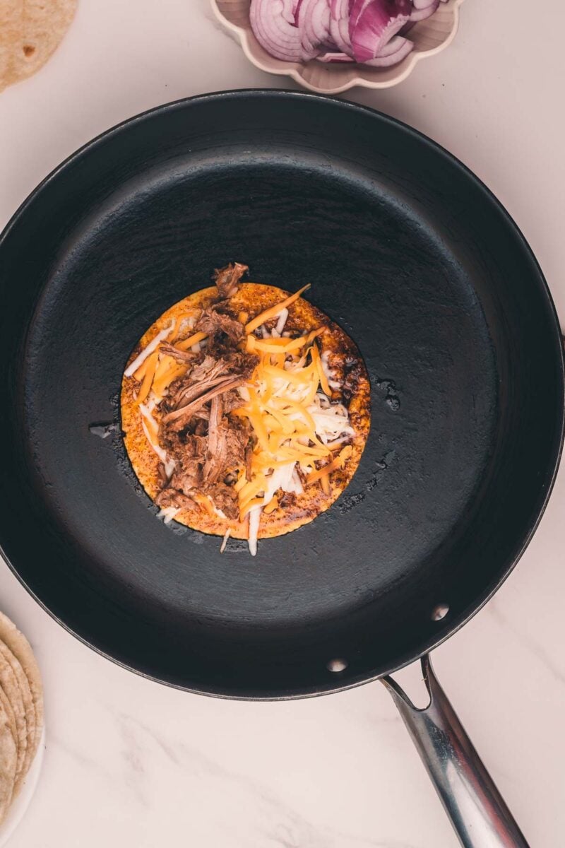 A corn tortilla topped with shredded meat and melted cheese is heating in a black skillet. Sliced red onions are in a bowl nearby.