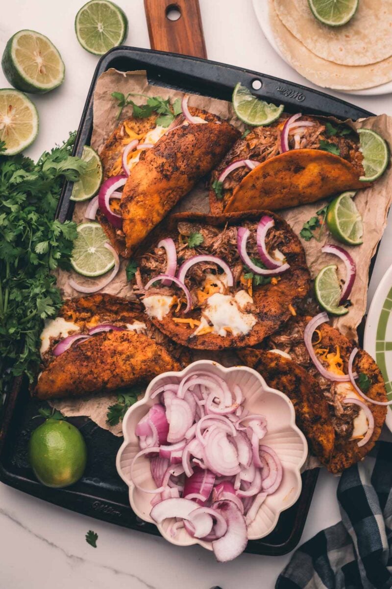 Tray of toasted shredded beef tacos with melted cheese, garnished with lime wedges and red onion slices, surrounded by fresh cilantro, lime halves, a bowl of sliced onions, and a stack of tortillas.