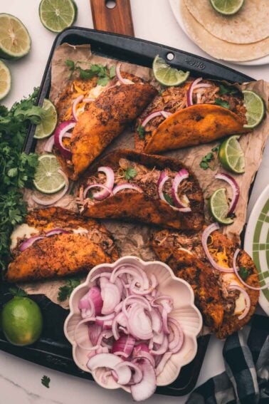 A tray of tacos filled with shredded meat, cheese, and onions, garnished with lime wedges and cilantro, with a bowl of sliced red onions and tortillas nearby.