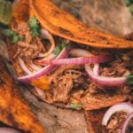 Close-up of tacos filled with shredded meat, melted cheese, red onion slices, and cilantro on parchment paper.