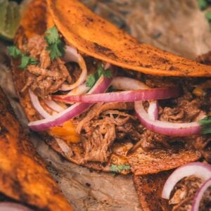 Close-up of tacos filled with shredded meat, melted cheese, red onion slices, and cilantro on parchment paper.