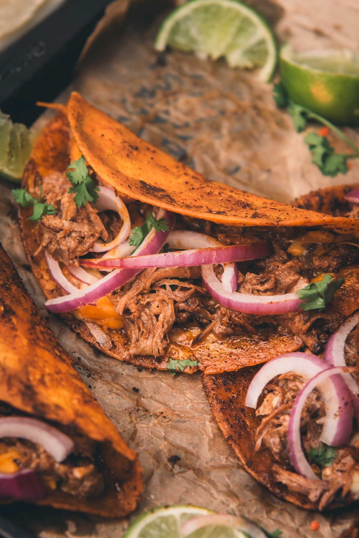 Three birria tacos filled with shredded meat, sliced red onions, and cilantro, served on parchment paper with lime wedges.