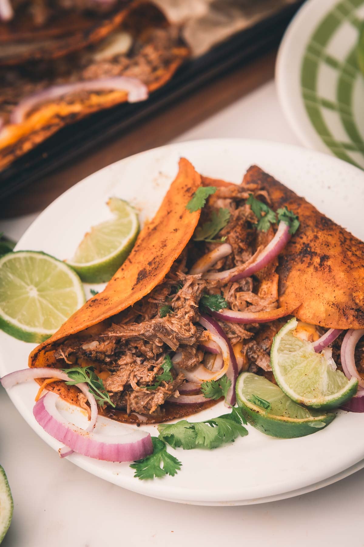 Two tacos with shredded mexican beef, red onion, cilantro, and lime wedges on a white plate; extra tacos and a green-patterned plate in the background.