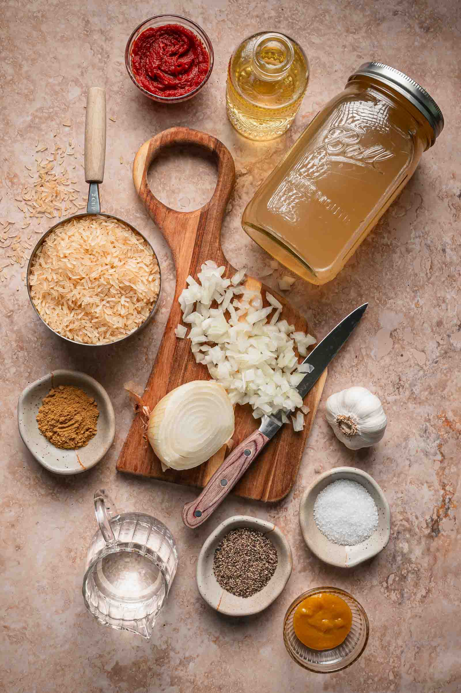 Chopped onion, garlic, spices, rice, tomato paste, broth, oil, and water are arranged on a countertop with a measuring cup, wooden board, and knife.