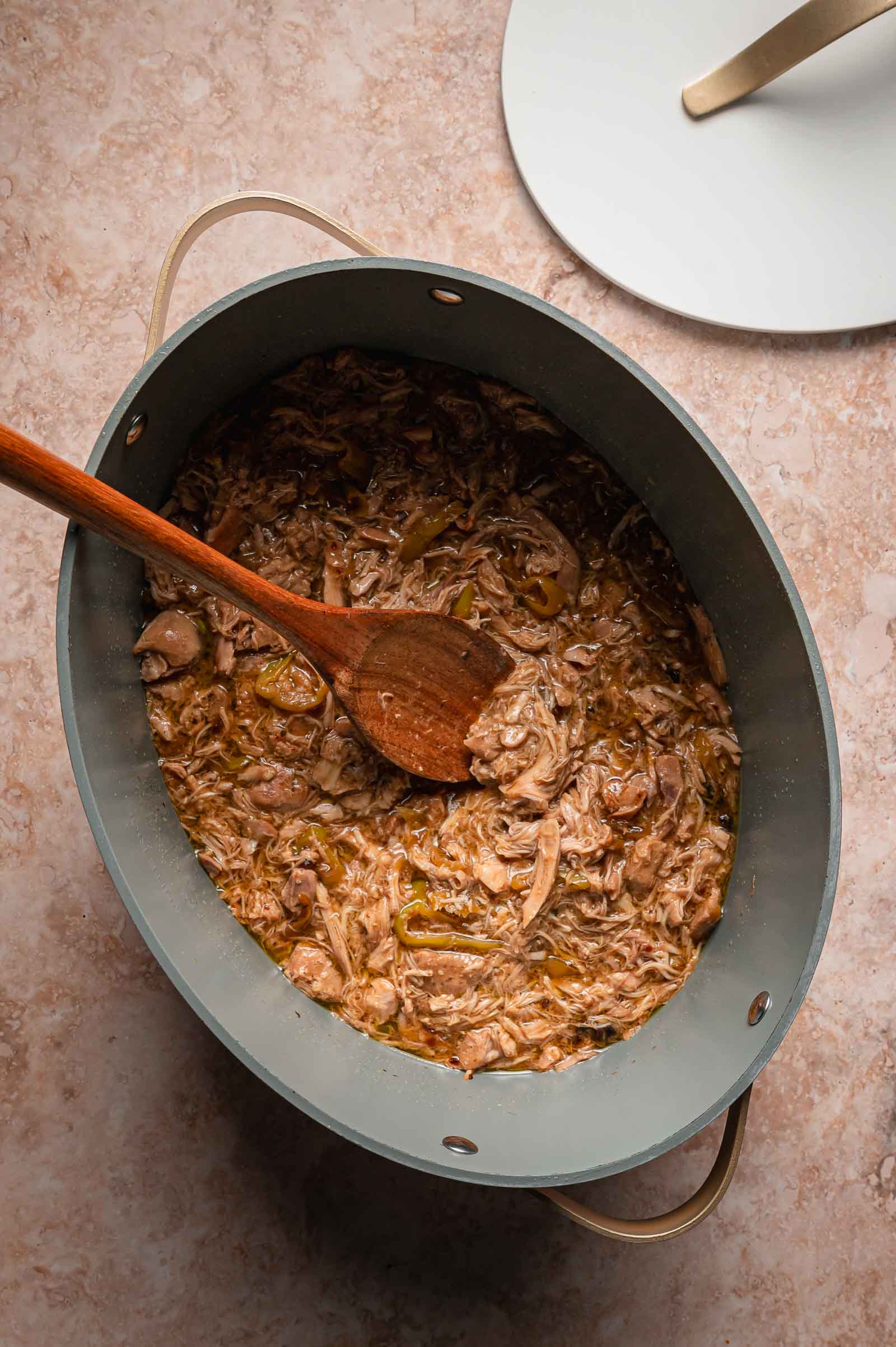 Oval pot filled with shredded Mississippi Chicken in sauce, stirred with a wooden spoon, on a light-colored countertop.