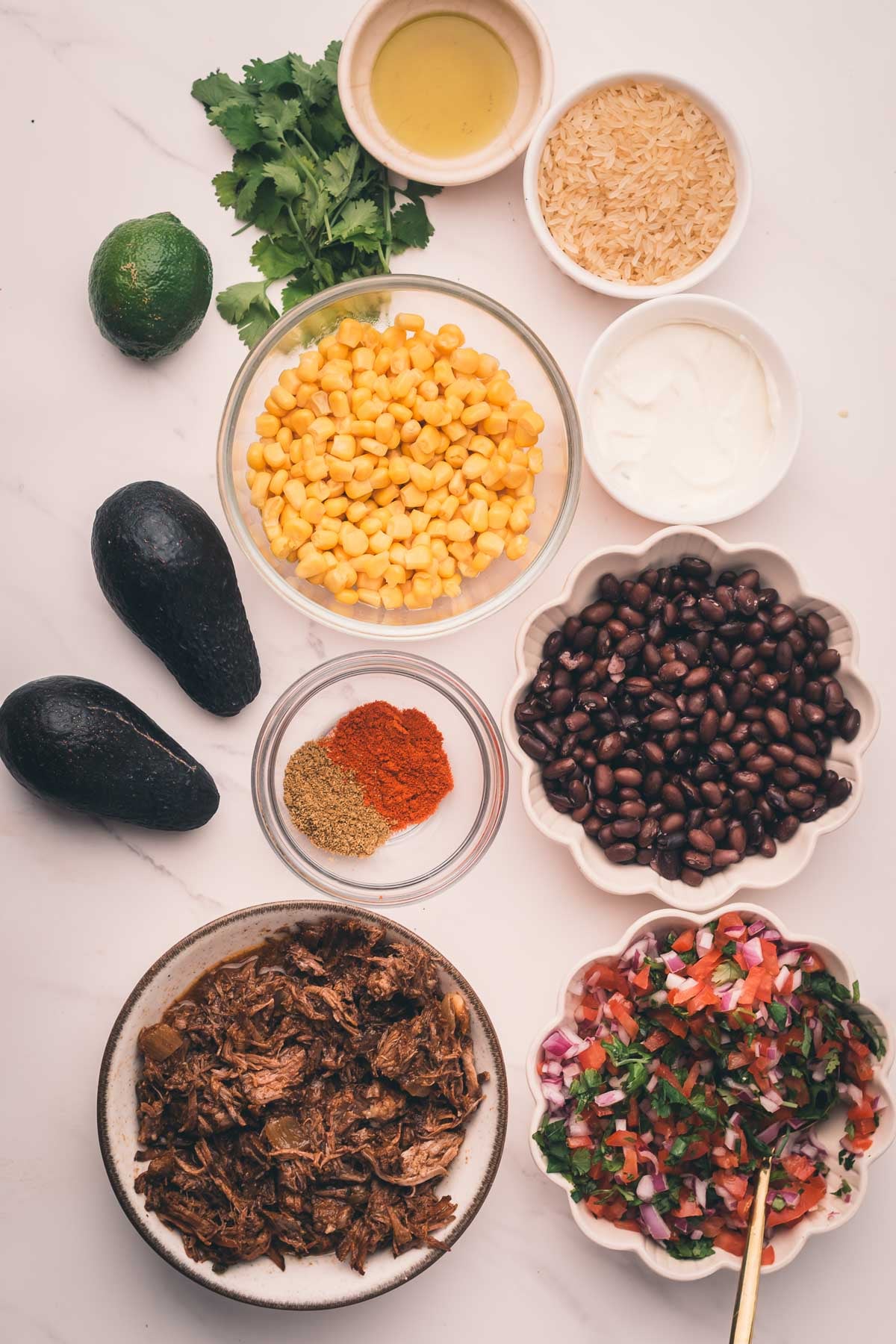 Ingredients for a burrito bowl arranged on a white surface: corn, black beans, rice, shredded meat, pico de gallo, avocado, sour cream, spices, lime, cilantro, and a small bowl of dressing.