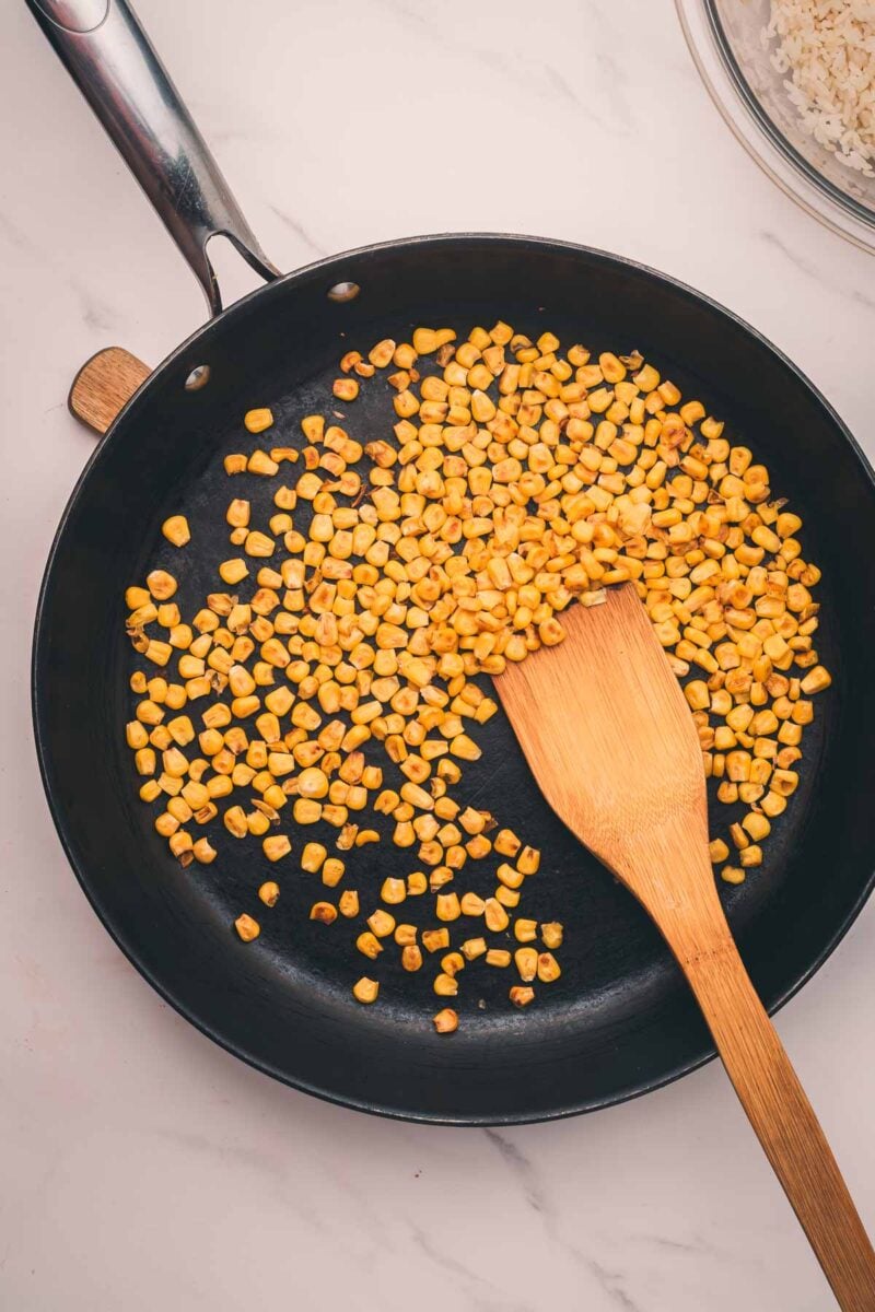 Corn kernels being sautéed in a black frying pan with a wooden spatula on a light marble surface.