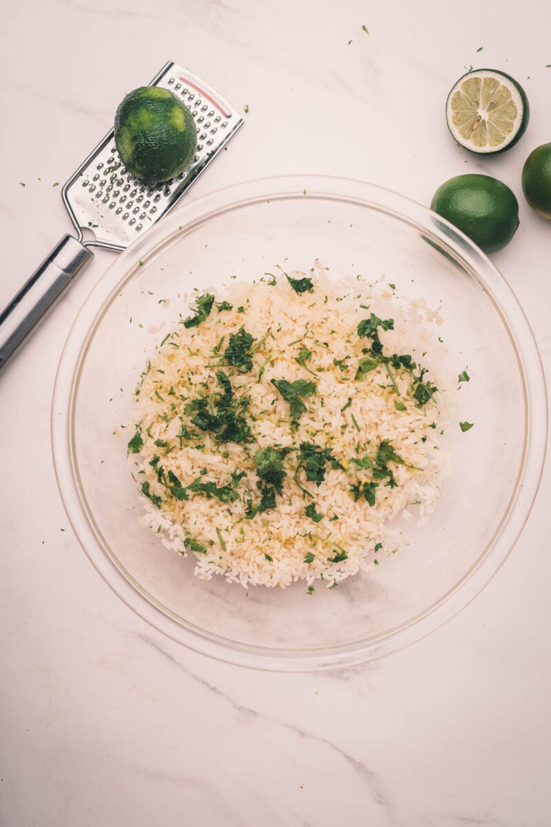 A glass bowl of cooked white rice with chopped cilantro and lime zest, next to a grater with a zested lime and two whole limes on a white surface.