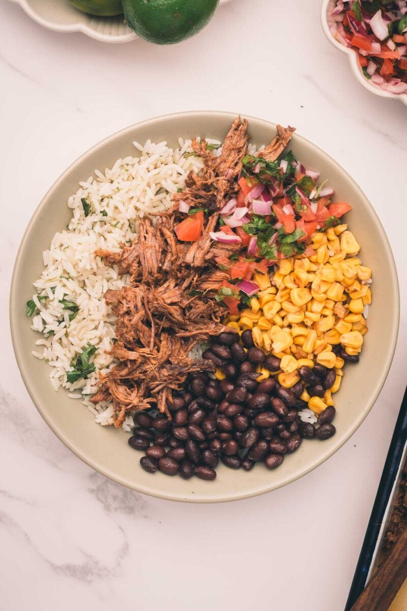 A bowl with shredded Mexican beef, white rice, black beans, corn, and fresh tomato salsa sits on a marble surface.