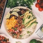 A bowl with shredded Mexican beef, sliced avocado, lime wedges, rice, black beans, corn, and pico de gallo, surrounded by fresh cilantro, tomatoes, limes, and other ingredients on a white surface.