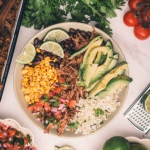 A bowl with shredded Mexican beef, sliced avocado, lime wedges, rice, black beans, corn, and pico de gallo, surrounded by fresh cilantro, tomatoes, limes, and other ingredients on a white surface.