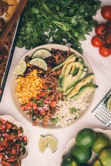 A bowl with shredded Mexican beef, sliced avocado, lime wedges, rice, black beans, corn, and pico de gallo, surrounded by fresh cilantro, tomatoes, limes, and other ingredients on a white surface.