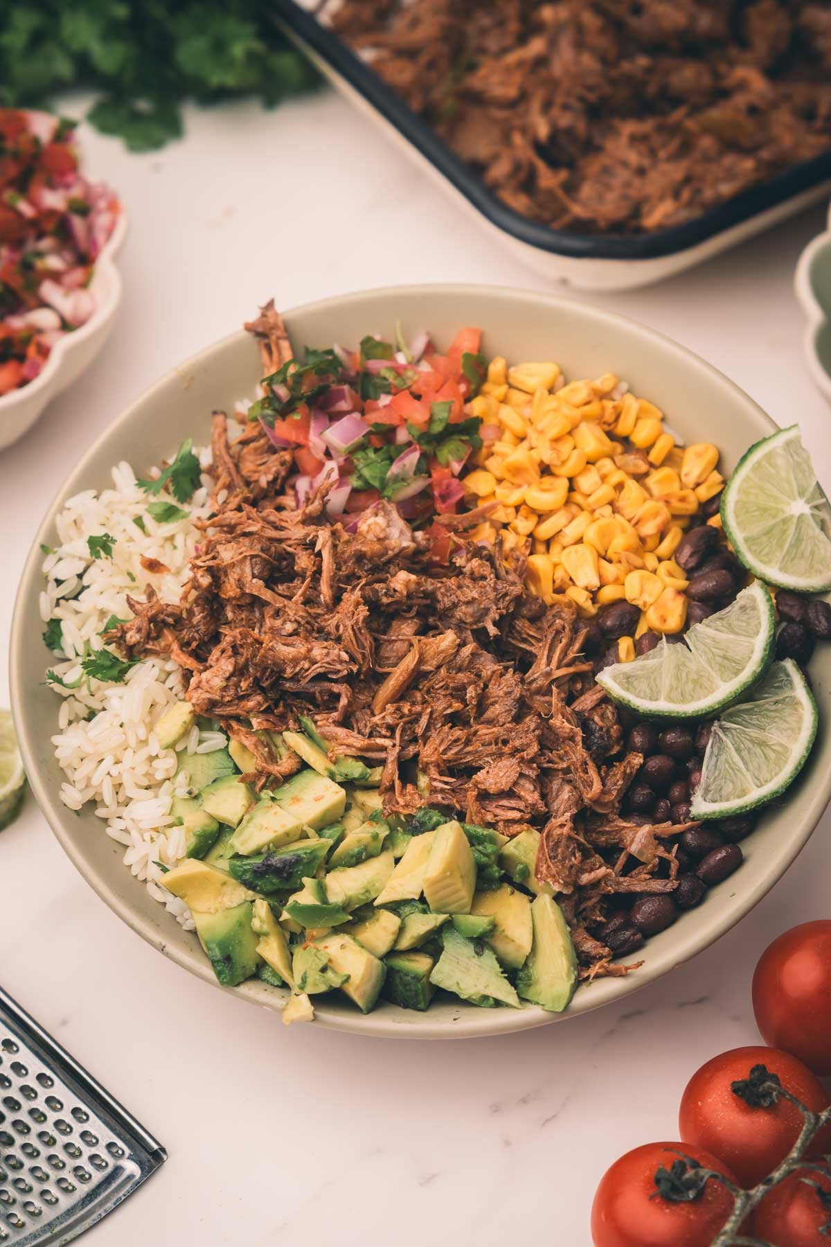 A bowl with shredded Mexican beef, white rice, avocado, corn, black beans, diced onions, cilantro, and lime wedges, arranged in sections.