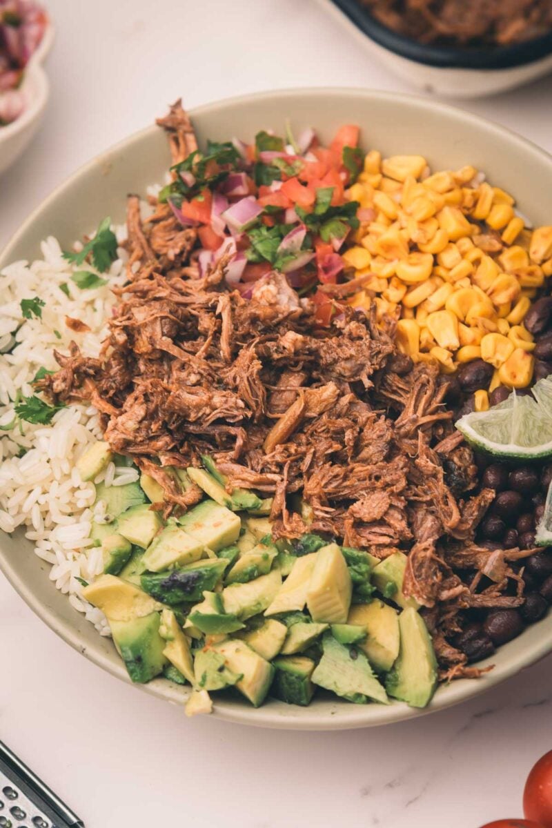 A bowl with shredded mexican beef, rice, black beans, corn, avocado, pico de gallo, and a lime wedge.