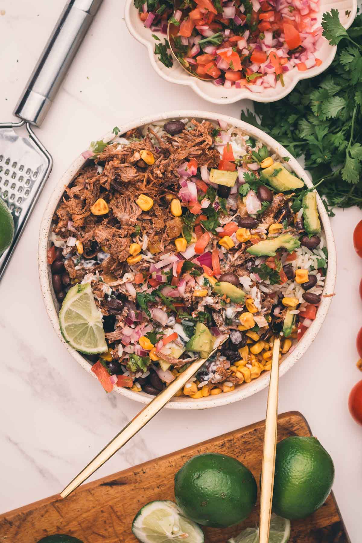 A bowl of shredded Mexican beef, corn, black beans, avocado, diced onions, and salsa with lime wedges, surrounded by fresh limes, cilantro, and a bowl of salsa on a white surface.
