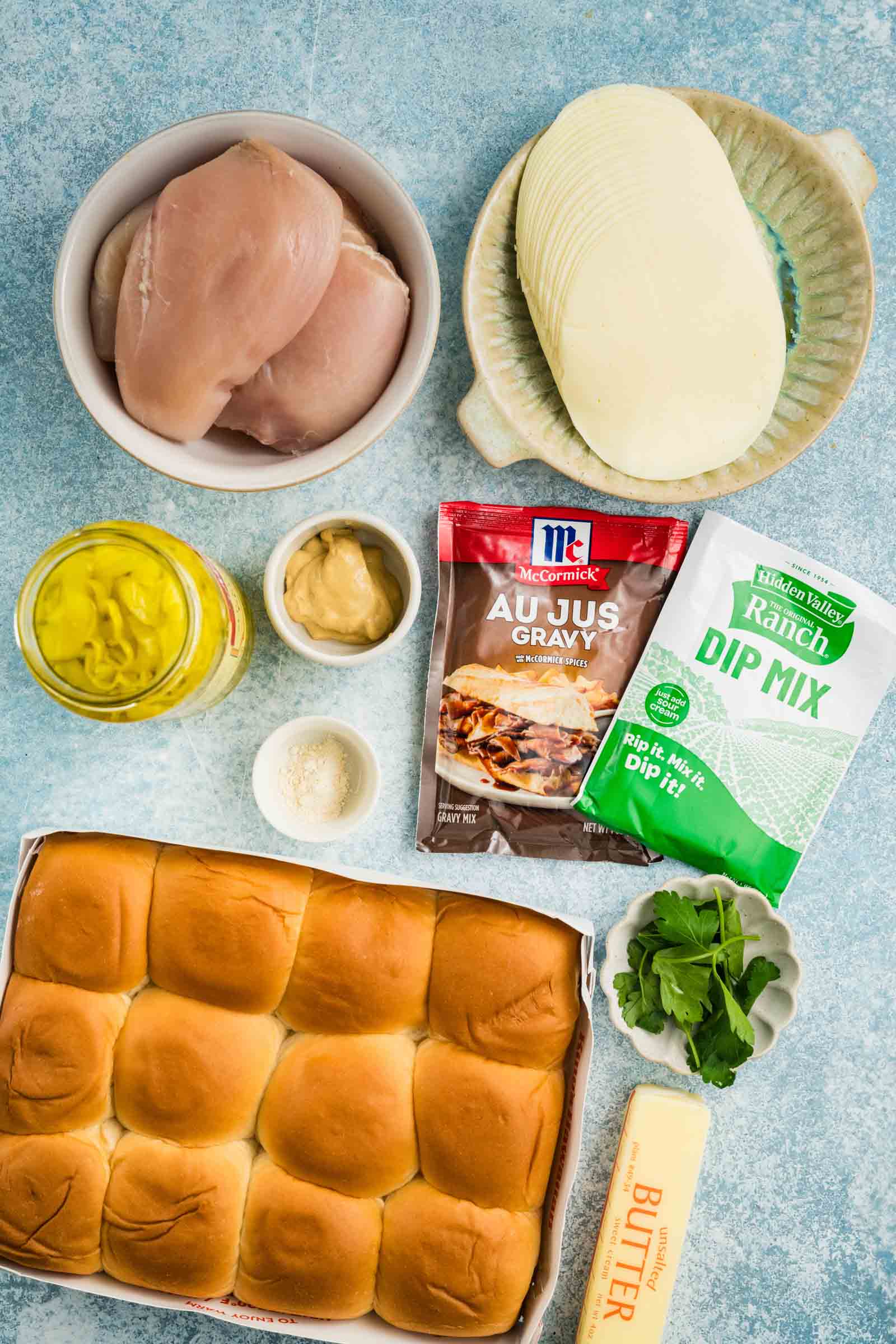 Ingredients on a blue surface: raw chicken breasts, provolone cheese, yellow mustard, mustard sauce, ranch dip mix, au jus gravy mix, butter, seasonings, parsley, and slider rolls.