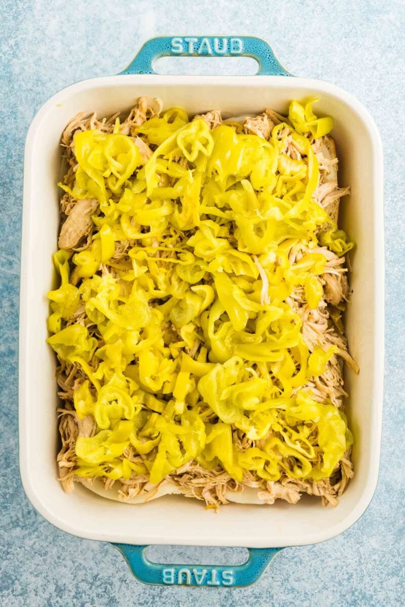 Rectangular blue Staub baking dish filled with shredded chicken topped with sliced yellow pepperoncini peppers on a light blue surface.