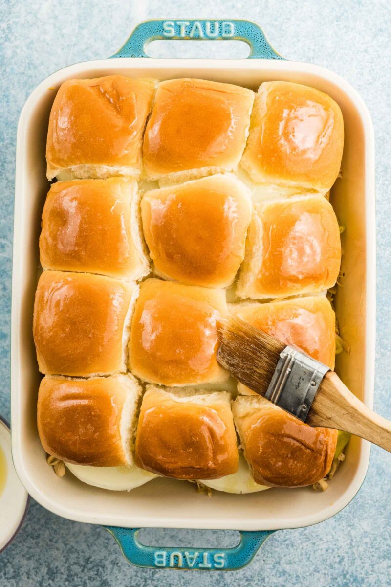 A baking dish with twelve golden slider rolls, some being brushed with melted butter using a pastry brush.