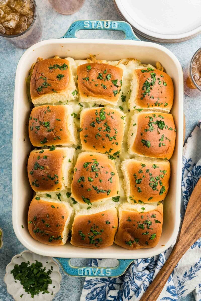 A baking dish filled with Mississippi chicken sliders topped with chopped herbs, placed on a blue surface with drinks and utensils nearby.
