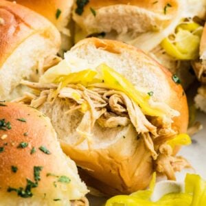 Close-up of shredded chicken and sliced pepperoncinis in soft slider rolls, topped with herbs, with extra peppers on the side.