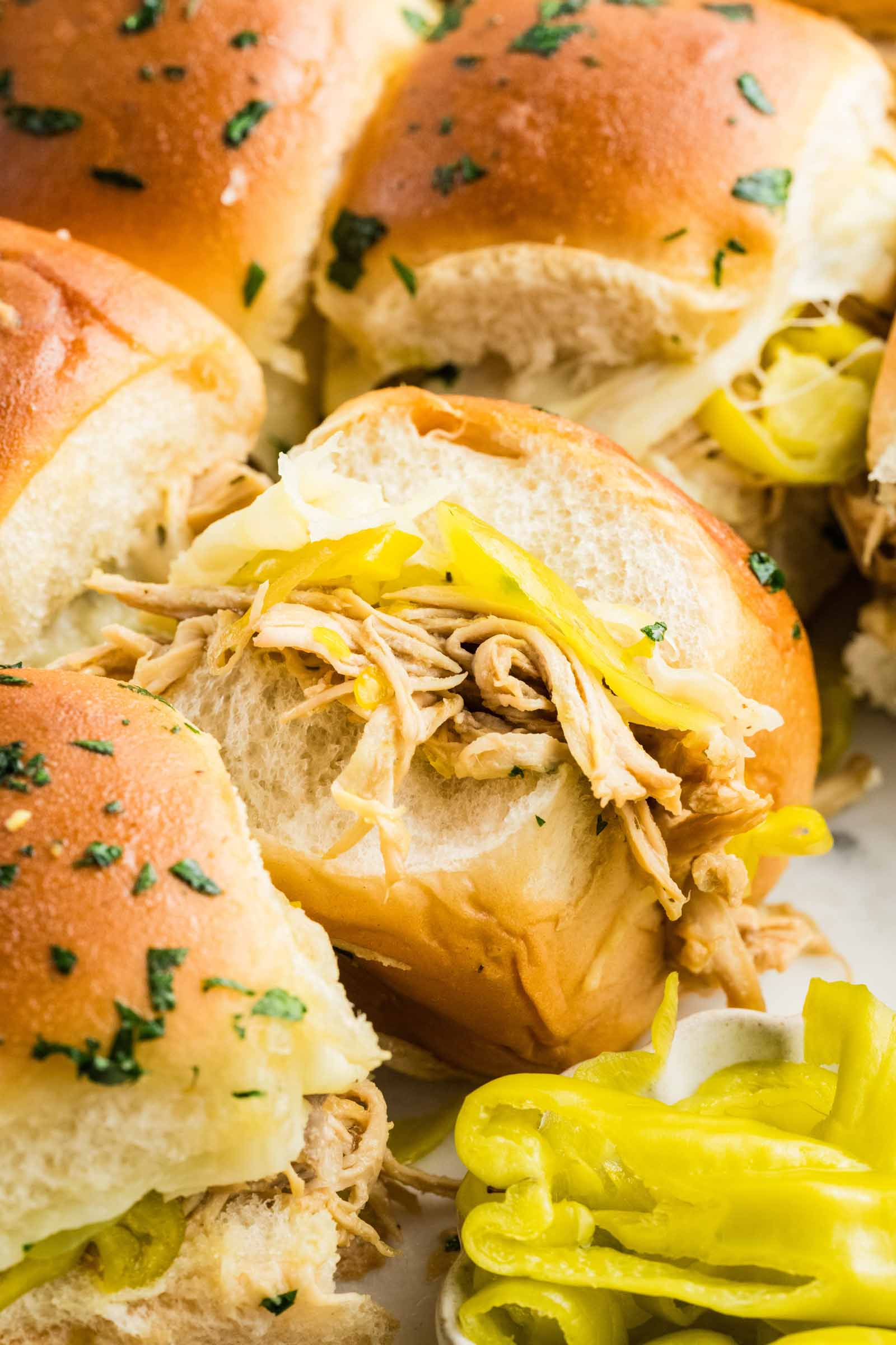 Close-up of shredded chicken and sliced pepperoncinis in soft slider rolls, topped with herbs, with extra peppers on the side.