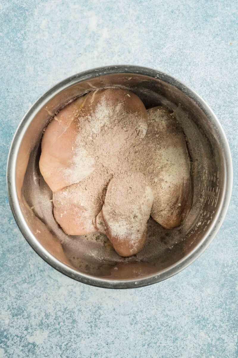 Raw chicken breasts in a metal bowl topped with a au just packet mix and ranch seasoning, set on a light blue textured surface.