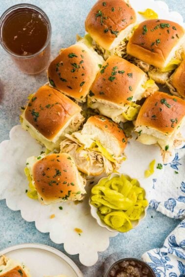 A platter of Mississippi chicken sliders on soft rolls with melted cheese and herbs, served with sliced pepperoncini and cold drinks on a blue and white table setting.