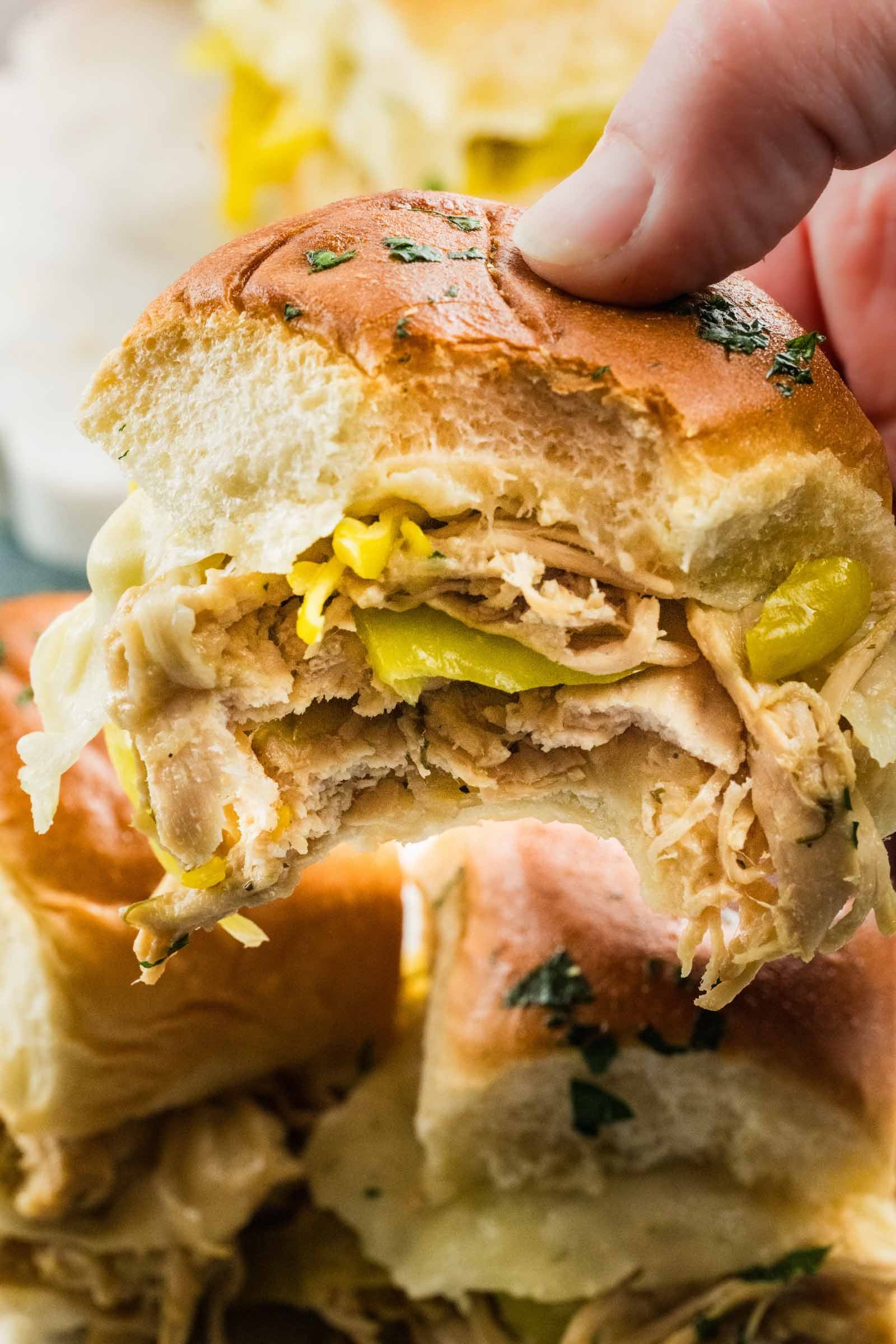 A close-up of a hand holding a Mississippi chicken slider with melted cheese and sliced pepperoncini on a soft bun, with more sandwiches in the background.