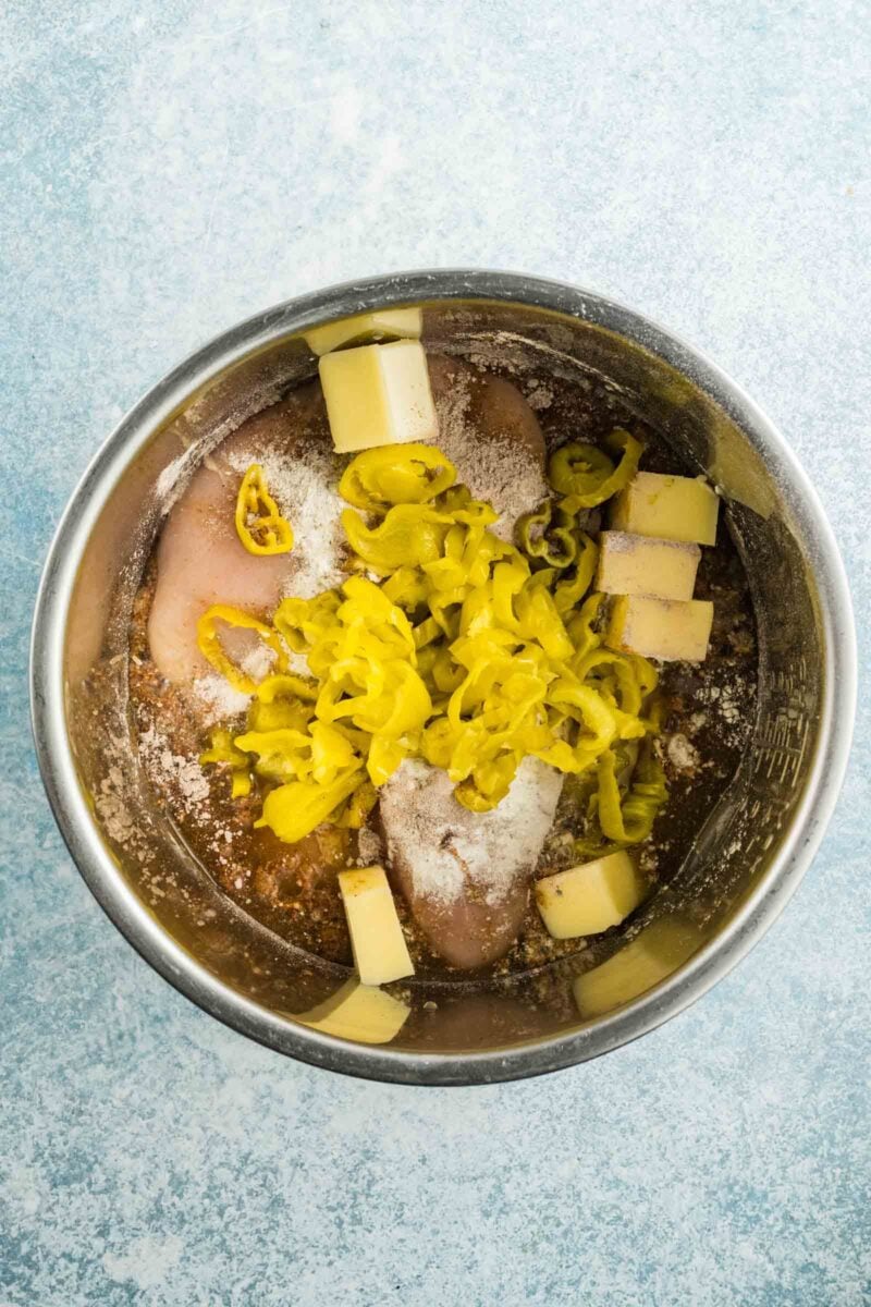 Raw chicken breasts in a metal bowl with butter slices, sliced yellow peppers, and seasoning on top, ready to be cooked.