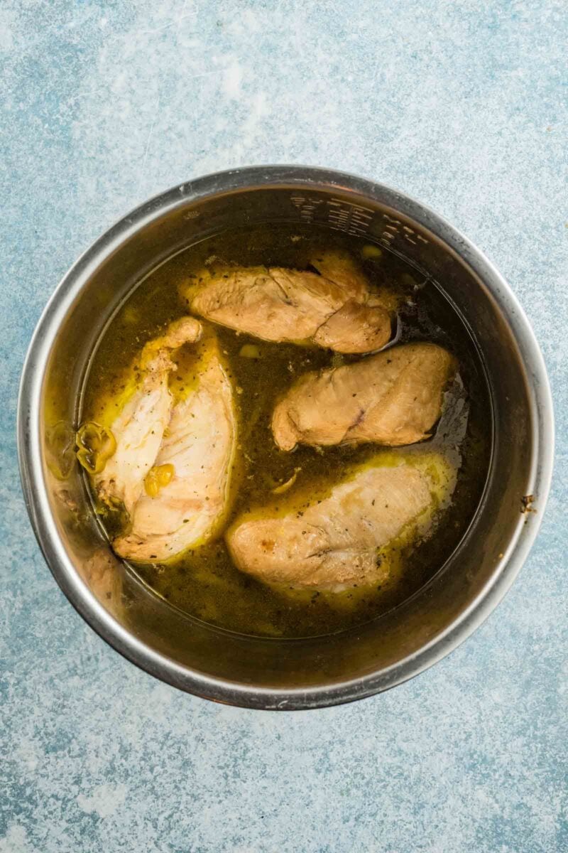 Four cooked chicken breasts in the pot of a pressure cooker with seasoned broth, viewed from above on a light blue textured surface.
