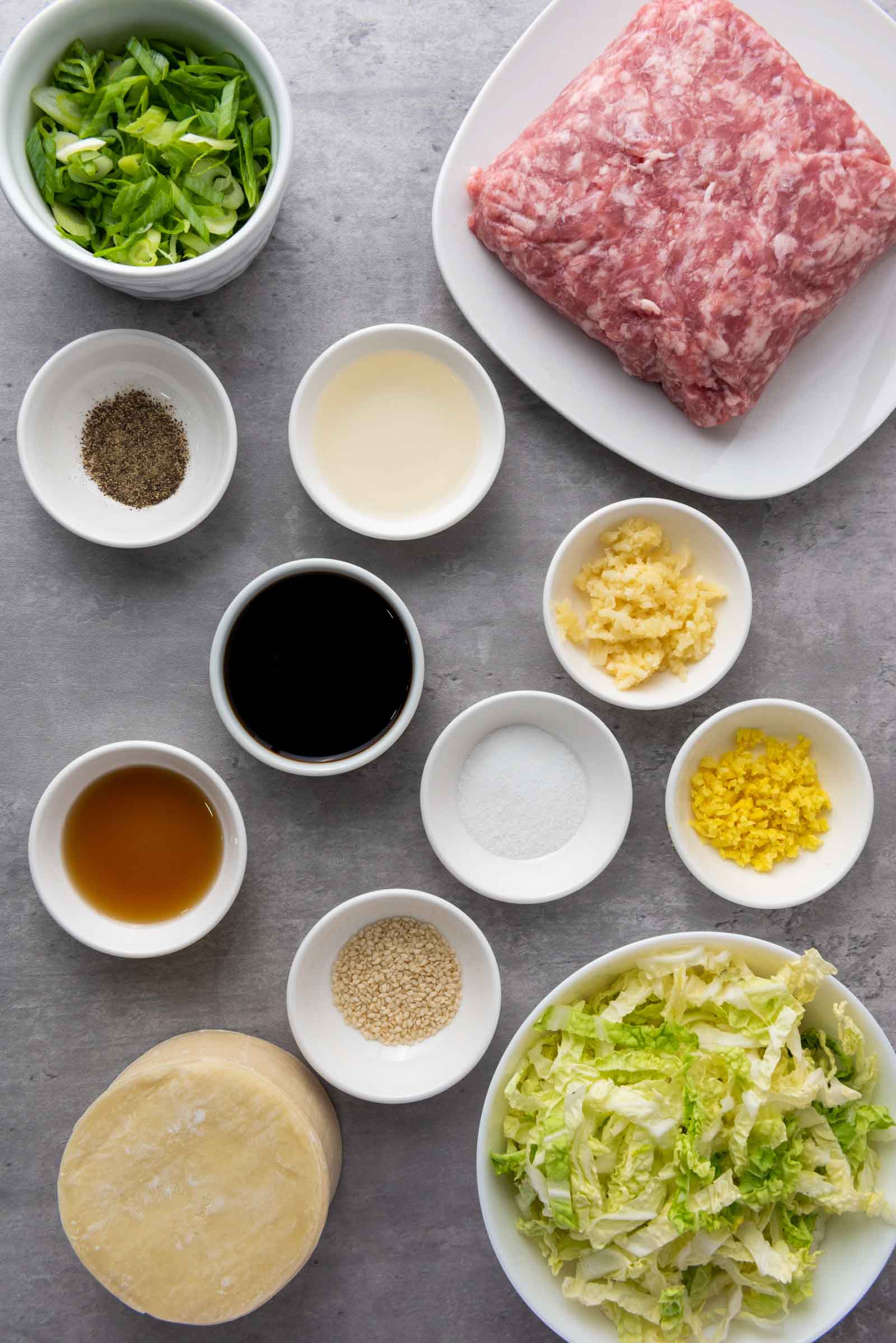 Ingredients for pork dumplings arranged on a gray surface: ground pork, chopped green onions, shredded cabbage, wrapper dough, soy sauce, sesame oil, garlic, ginger, sugar, sesame seeds, and pepper.